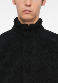 Svart fleecejakke med høy krage og myk tekstur, med glidelås i halsen og en tettsittende design.