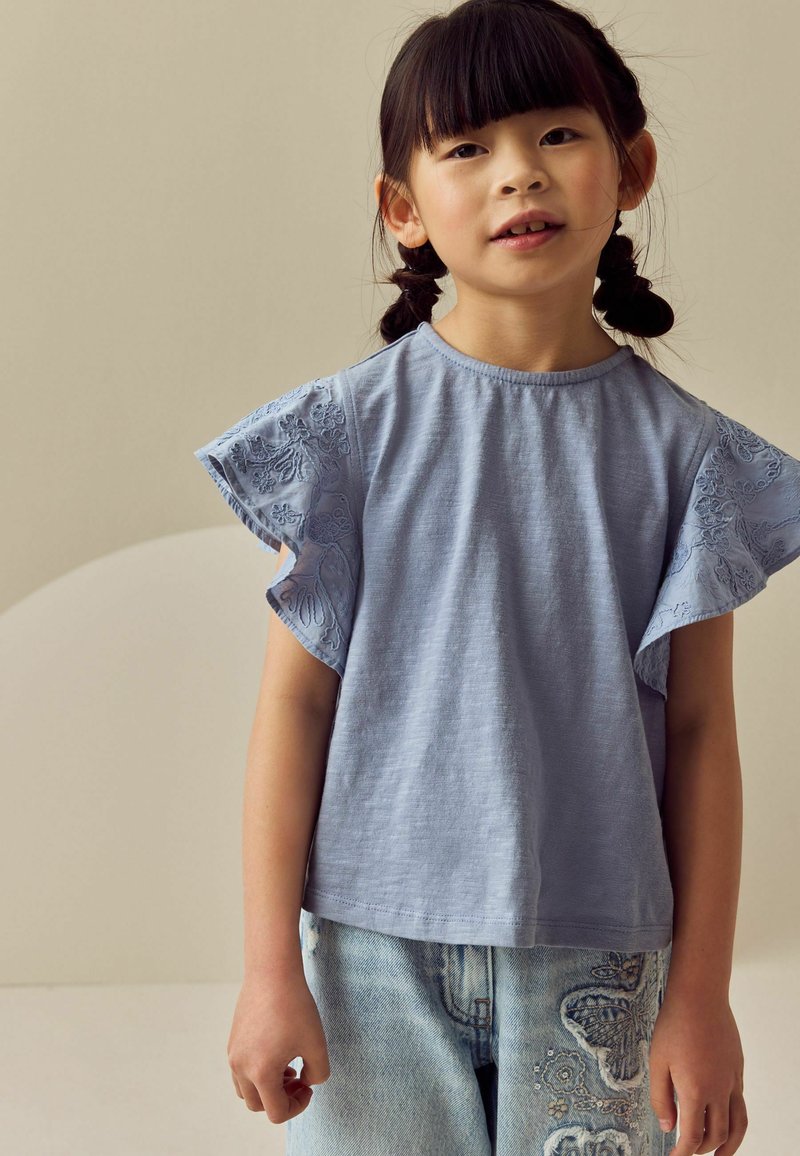 Jeune fille aux cheveux tressés portant un top bleu clair brodé à manches courtes et un jean en denim clair avec des patchs floraux.