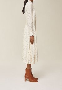 Kvinna i en krämfärgad blommig midiklänning och bruna klackade ankla stövlar stående i profil mot en neutral bakgrund.