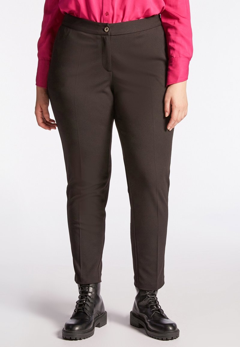 Personne portant un pantalon ajusté marron foncé, des bottes noires à lacets et une chemise rose vif à manches longues, debout devant un fond blanc.