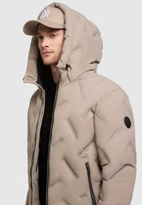 Giubbotto imbottito beige con design a rilievo, grande cappuccio, zip nera e tasche laterali. Decorato con un'etichetta rotonda sul maniche sinistro.