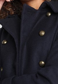Cappotto in lana navy con design doppiopetto, grandi bottoni in ottone e colletto strutturato. Texture morbida con linee pulite.