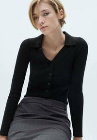 Zwarte geribbelde lange mouwen cardigan met een kraag, voorzien van een button-front en een slim fit. Draagt bij een grijze gestreepte rok.
