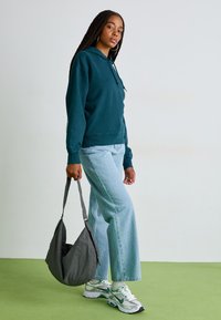 Sweatshirt à capuche bleu-vert avec une poche frontale de type kangourou, associé à un jean large bleu clair et un sac à épaule texturé gris. Baskets blanches.