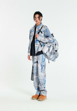 Joven con chaqueta y pantalones a juego con patrón azul y blanco, llevando una mochila grande con el mismo diseño y zapatos color canela.