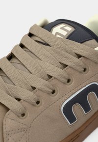 Tan suede sneaker with textured surface, beige laces, and a dark blue accent on the tongue. Features a prominent white logo on the side.