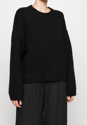 Persoon die een losse zwarte, pluizige trui met lange mouwen en zwarte wijdvallende broek draagt tegen een effen achtergrond.