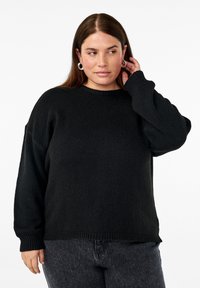 Zwarte gebreide trui met een ribbelstructuur, ronde hals en licht oversized mouwen. Gecombineerd met donkere jeans voor een casual uitstraling.