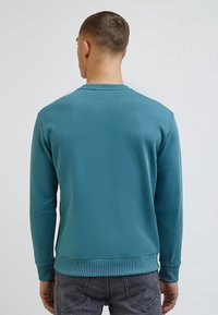 Tyrkysová mikina s kulatým výstřihem, dlouhými rukávy, žebrovanými manžetami a lemem. Látka působí hladce a lehce, vhodná pro běžné nošení.