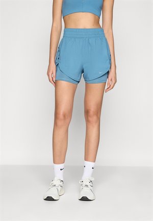Kvinde iført blå atletikshorts og crop top, hvide Nike-strømper og hvide sneakers, der står mod en ensartet lys baggrund.