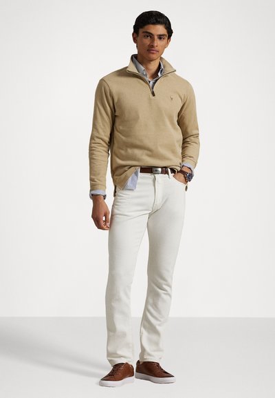 Beige zippered pullover made of soft fabric, paired with white jeans. Brown sneakers with white soles. Striped collared shirt visible underneath.
