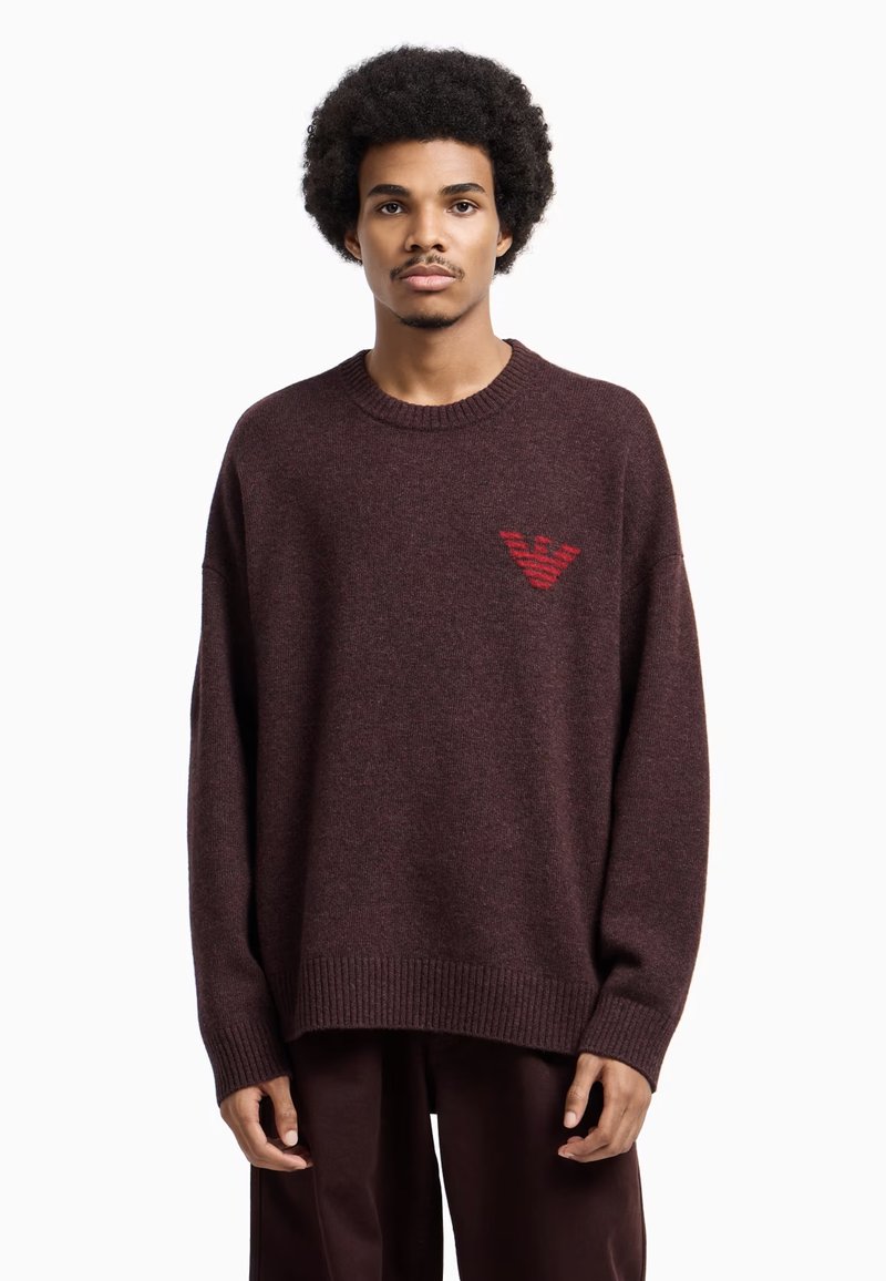 Maglione oversize color burgundy realizzato in tessuto morbido, con collo e polsini a coste, e un logo rosso sul lato sinistro del petto. Design semplice.