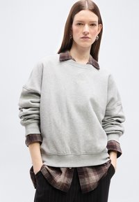 Jeune femme aux cheveux longs et raides portant un sweat-shirt gris clair sur une chemise à carreaux marron et un pantalon noir à fines rayures, les mains dans les poches.