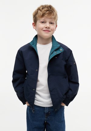 Ragazzo giovane con le mani nelle tasche, indossa una giacca blu scuro sopra una camicia bianca e jeans blu, sorridente su uno sfondo semplice.