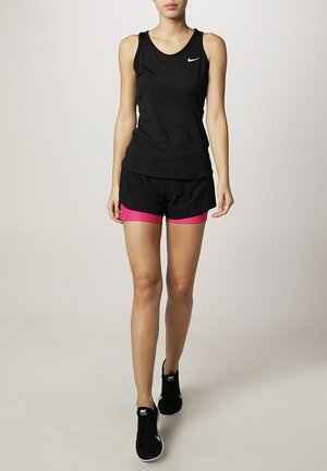 Femme marchant portant un débardeur noir Nike, un short noir avec une doublure rose et des baskets noires Nike sur un fond uni.