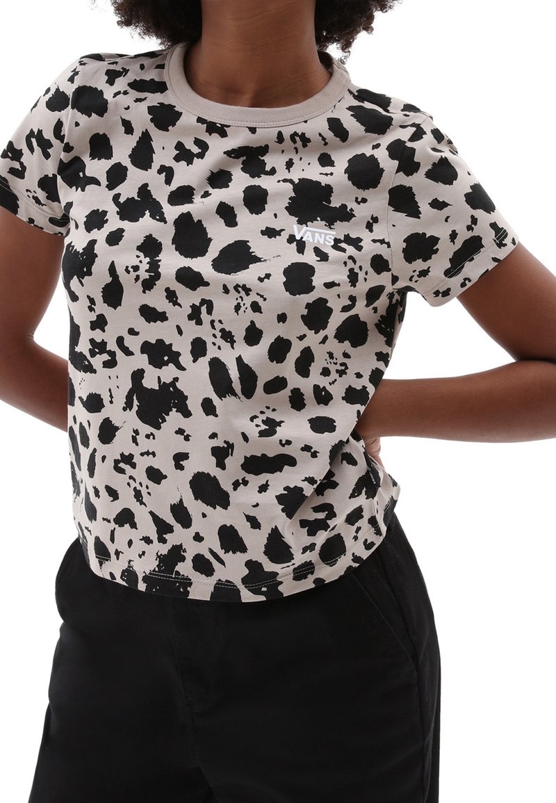 Personne portant un t-shirt Vans beige à manches courtes avec des taches noires, les mains sur les hanches, associé à un pantalon noir.