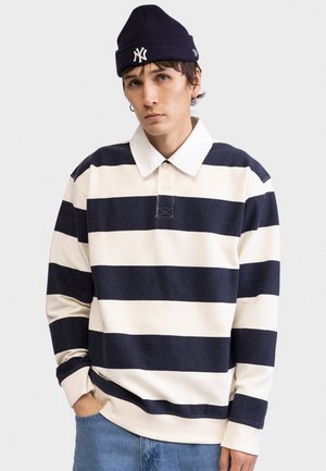 Polo a maniche lunghe a righe nei colori blu navy e crema, con texture a coste e colletto classico. Indossata con un berretto a maglia blu navy.