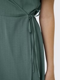 Primo piano di un vestito avvolgente verde legato a fiocco in vita, che mostra un tessuto strutturato e un braccio nudo accanto.