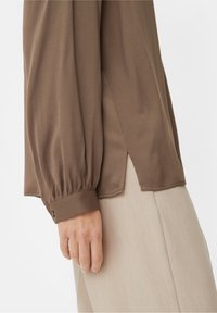 Blouse marron à manches longues avec poignets froncés, fentes latérales, texture lisse et coupe décontractée, associée à un pantalon beige clair.