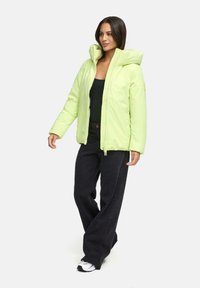 Vrouw in een lichtgroene gewatteerde jas met kap, zwarte top, wijde zwarte broek en witte sneakers, staand tegen een effen achtergrond.