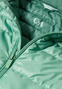 Interior de chaqueta acolchada verde ligera con textura acolchada, que cuenta con una cremallera y etiqueta de C&A con detalles del material de la tela.