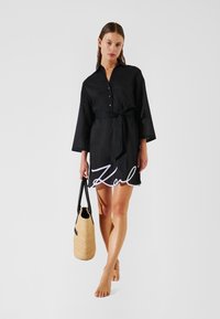 Vestido camisa negro con botones, cintura anudada, mangas tres cuartos y un dobladillo con forma de ondas que presenta un logo blanco. Combinado con un bolso trenzado color beige.