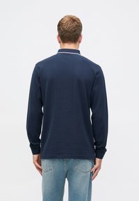 Tumši zils garām piedurknēm polo krekls ar kontrastējošu balto apkakli, pieguļošs, izgatavots no mīkstas kokvilnas auduma, parādīts uz modeļa no aizmugures.