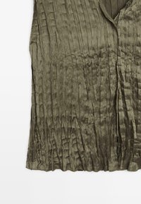 Top verde oliva senza maniche realizzato in tessuto strutturato, con pieghe verticali e scollo a V con bordi grezzi.