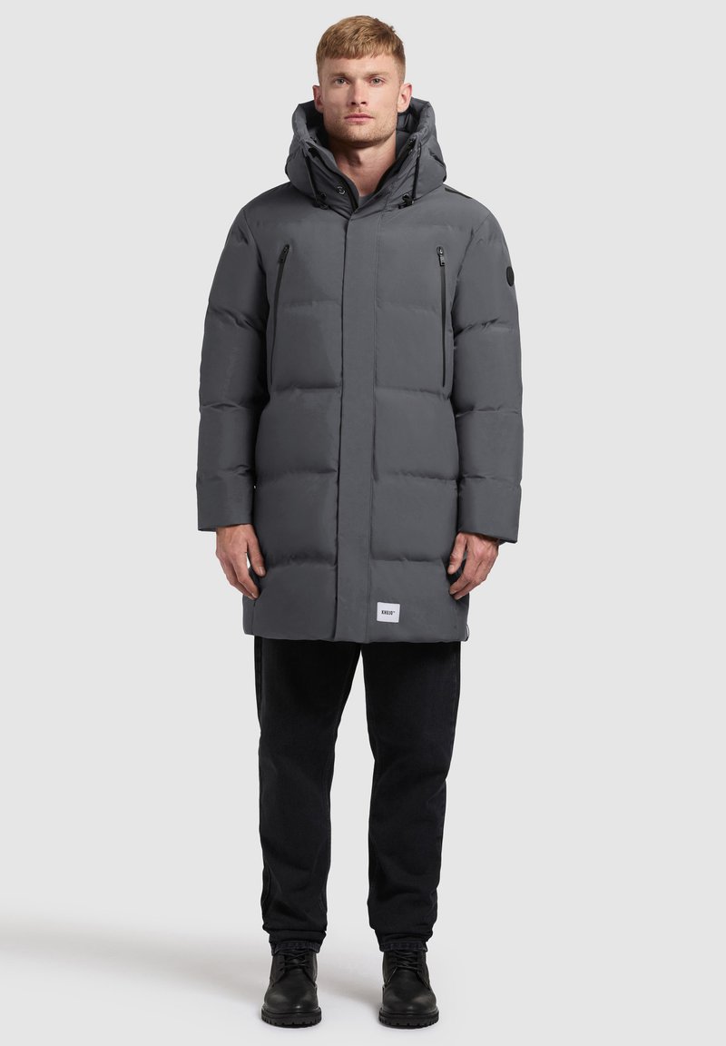 Muškarac stoji odjeven u dugi sivi zimski kaput s kapuljačom, crne hlače i crne čizme na jednostavnoj svijetloj pozadini.