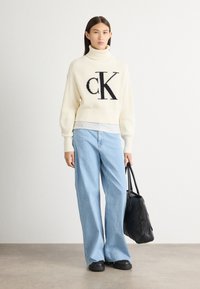 Dicker cremefarbener Rollkragenpullover mit schwarzem "CK"-Logo, kombiniert mit hellblauen Schlagjeans. Schwarze strukturierte Tragetasche und dunkle Schuhe.