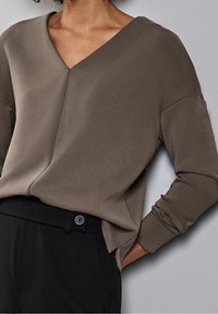 V-ringad långärmad topp i dämpad taupe-färgad tyg, med en avslappnad passform och jämn textur, tillsammans med svarta högmidjade byxor.