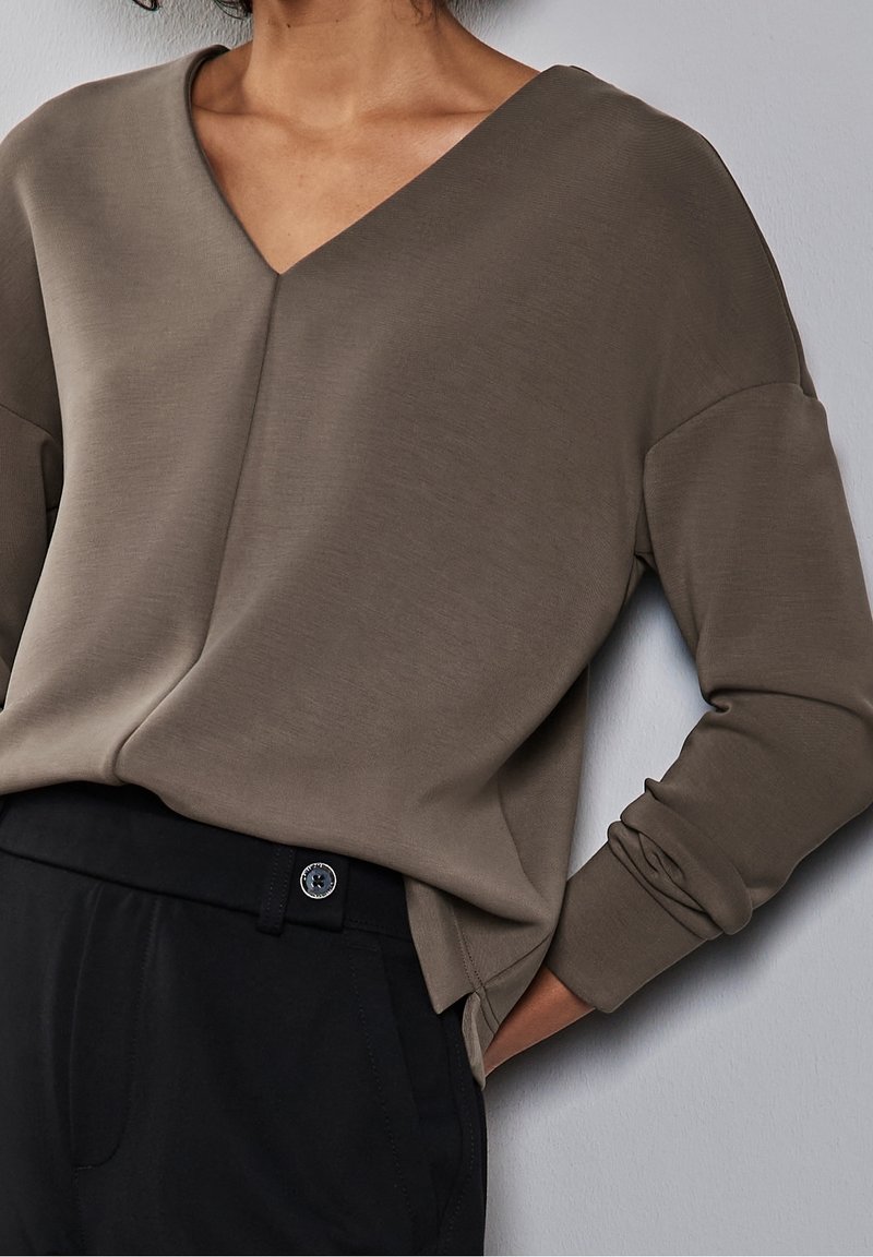 V-ringad långärmad topp i dämpad taupe-färgad tyg, med en avslappnad passform och jämn textur, tillsammans med svarta högmidjade byxor.