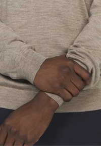 Jasnobrązowy sweter o fakturze z prążkowanymi mankietami, noszony z założonymi skrzyżowanymi ramionami. Tkanina ma subtelny melanżowy wzór.