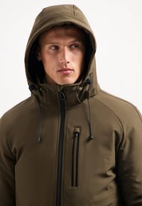 Olijfgroene geïsoleerde jas met een capuchon, een rits aan de voorkant, zijzakken en verstelbare trekkoorden voor een goede pasvorm en warmte.