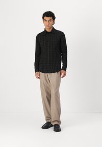 Camisa a cuadros negro y verde oliva con botones al frente, de manga larga, combinada con pantalones beige y zapatos negros. Diseño simple y texturizado.