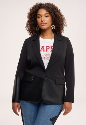 Kvinde med krøllet hår bærer stjerneøreringe, sort blazer med blandede teksturer, hvid grafisk skjorte og blå jeans med sort blondedetalje.