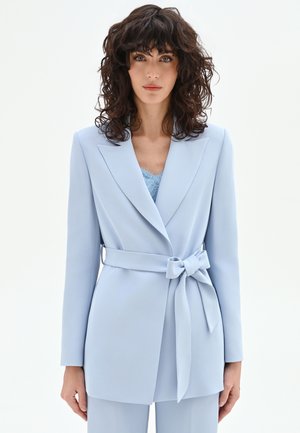 Femme aux cheveux bouclés et foncés portant un blazer bleu clair ceinturé sur un haut en dentelle, debout devant un fond blanc uni.