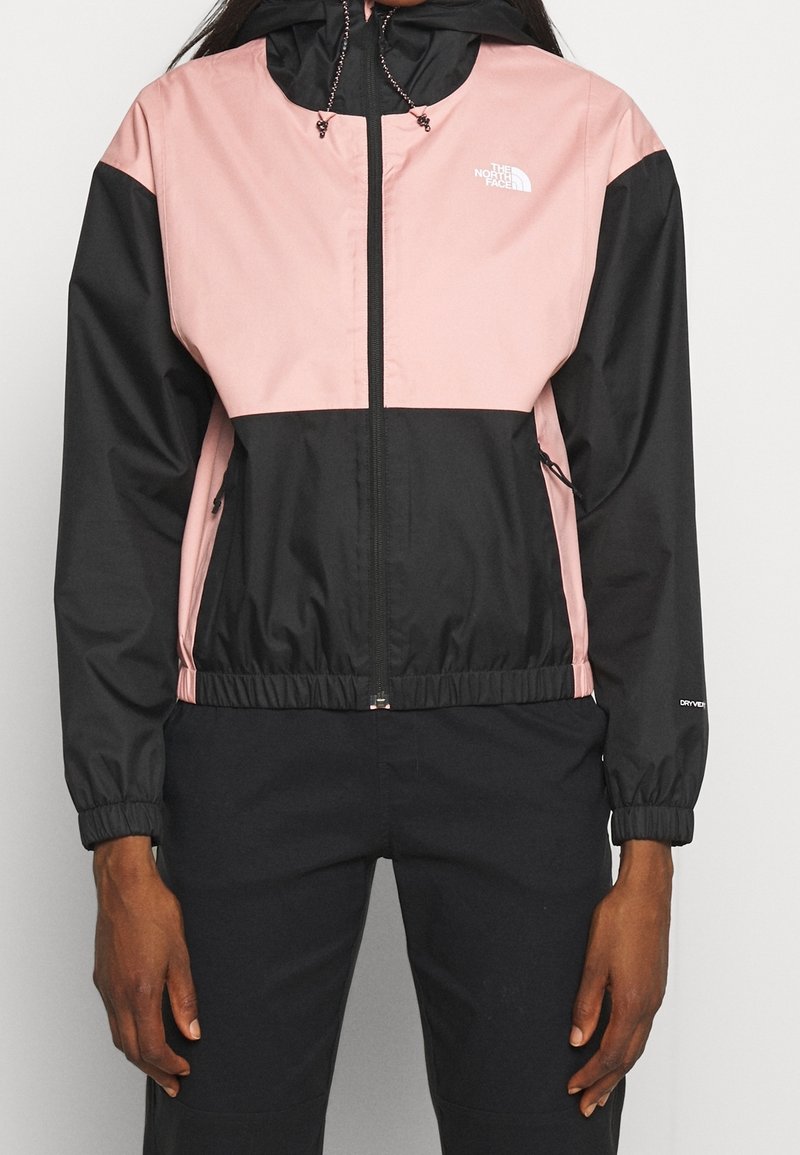Personne portant une veste à capuche North Face rose clair et noire avec fermeture éclair sur le devant et un pantalon noir, debout les bras détendus le long du corps.