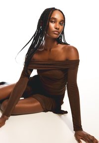 Abito marrone off-the-shoulder con maniche lunghe, tessuto testurizzato, design aderente e vita leggermente arricciata. La modella è seduta su una superficie chiara.