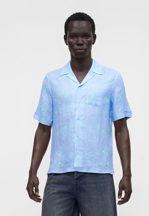 Homme portant une chemise bleu clair à manches courtes avec des motifs subtils de tortues et un jean bleu foncé sur un fond uni.