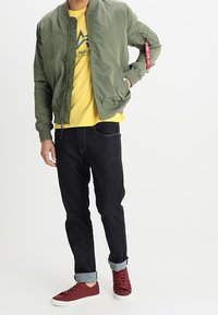 Olivgrön bomberjacka med ribbad krage, gul t-shirt, mörk denimjeans och vinröda sneakers med vita sulor och uppvikta ben.