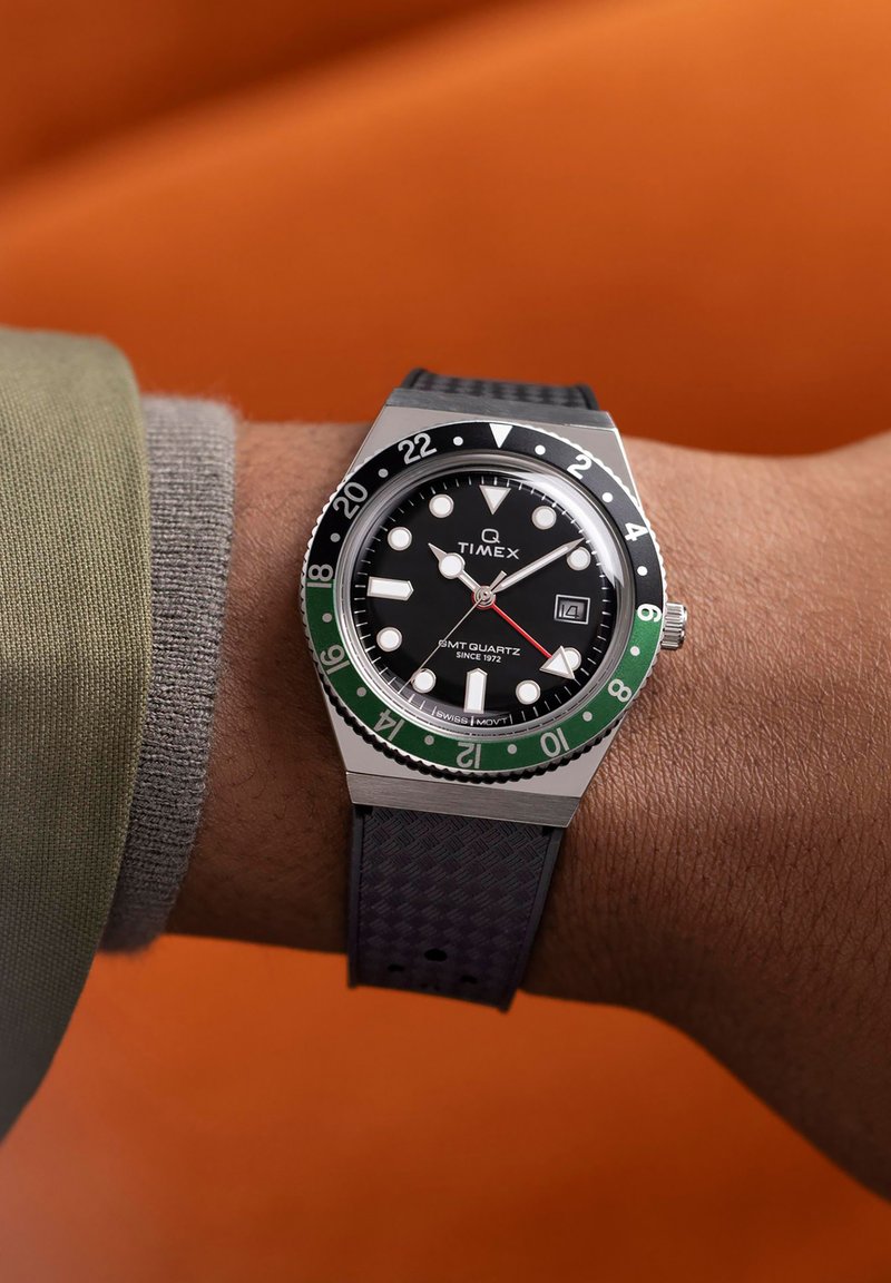 Montre Timex Q GMT avec boîtier en acier inoxydable, cadran noir, lunette verte et bracelet en caoutchouc noir texturé affichée sur un poignet.