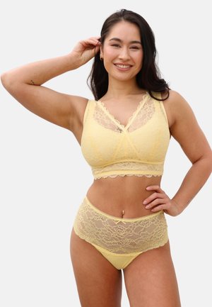 Woman smiling, wearing a yellow lace bralette and matching high-waisted lace underwear, posing with one hand on hip and the other touching her hair.