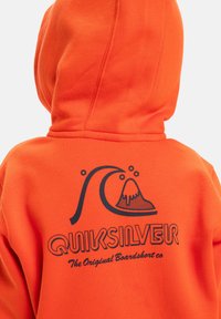 Haut à capuche orange avec un grand graphisme représentant des vagues et un volcan. Le dos affiche le texte "Quiksilver" en noir. Texture de tissu lisse.