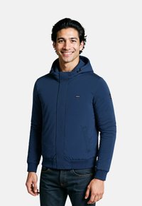 Marineblauer Pufferjacke mit Kapuze, Frontreißverschluss, gerippten Bündchen und zwei seitlichen Taschen. Glatte Textur mit minimalen Markenhinweisen.