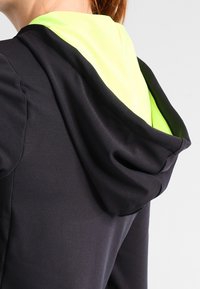 Gros plan sur une personne portant un sweat à capuche noir avec une doublure de capuche jaune néon, vu de l'arrière et de la zone de l'épaule.