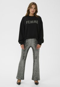 Naine seisab seljas musta dressipluusi, millel on tekst "FEMME", ning hõbedaste sequinsärkidega laienevad püksid, mida täiendab mustade teravate kontsadega kingad.
