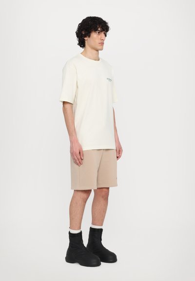 T-shirt en coton beige clair à manches courtes avec un petit motif graphique vert. Associé à un short beige et des bottines noires à semelles épaisses.