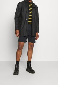 Černá voděodolná bunda s kapsami na zip, žlutě pruhované tričko, tmavé šortky a černé kotníkové boty s texturovanou podrážkou.