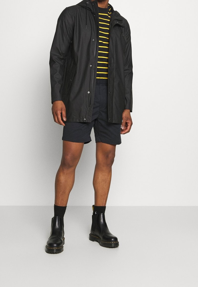 Černá voděodolná bunda s kapsami na zip, žlutě pruhované tričko, tmavé šortky a černé kotníkové boty s texturovanou podrážkou.